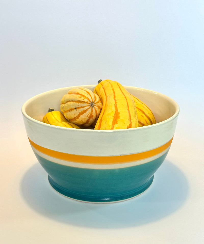 A ceramic bowl with a white top half, a yellow-orange stripe, and a teal-blue bottom half, filled with decorative gourds and small pumpkins on a light gray background.