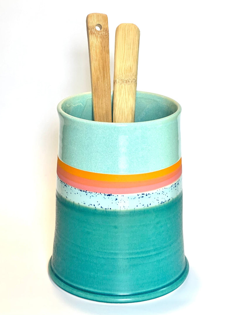 A ceramic kitchen utensil holder in a mint green and teal glaze with a retro orange and pink stripe, containing two wooden utensils, on a white background.